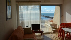 A room with a sofa, a TV, and a table with chairs, overlooking a beach with the ocean and rocky shore.