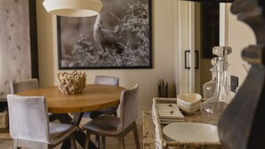 A dining area with a wooden table, chairs, a hanging light fixture, a framed picture on the wall, and a wicker side table with glassware.