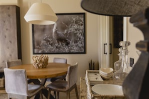 A dining area with a wooden table, chairs, a hanging light fixture, a framed picture on the wall, and a wicker side table with glassware.