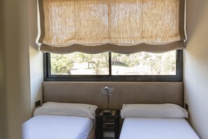 A room with two beds, a window with a view of trees, and a small bedside table with a lamp.