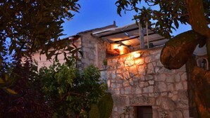 A stone building with a warm light source near the entrance, surrounded by trees and shrubs. - Apanemia Stone House in Crete (Apokoronas)