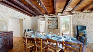 Un comedor rústico con una mesa de madera dispuesta para seis personas, rodeada de sillas de madera. Se observa una pared de piedra y una chimenea al fondo.