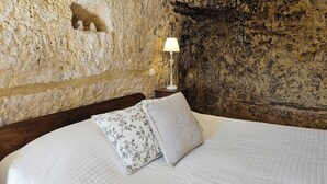 Una cama con ropa de cama blanca y almohadas florales, una mesita de noche con lámpara, y una pared de piedra texturizada.