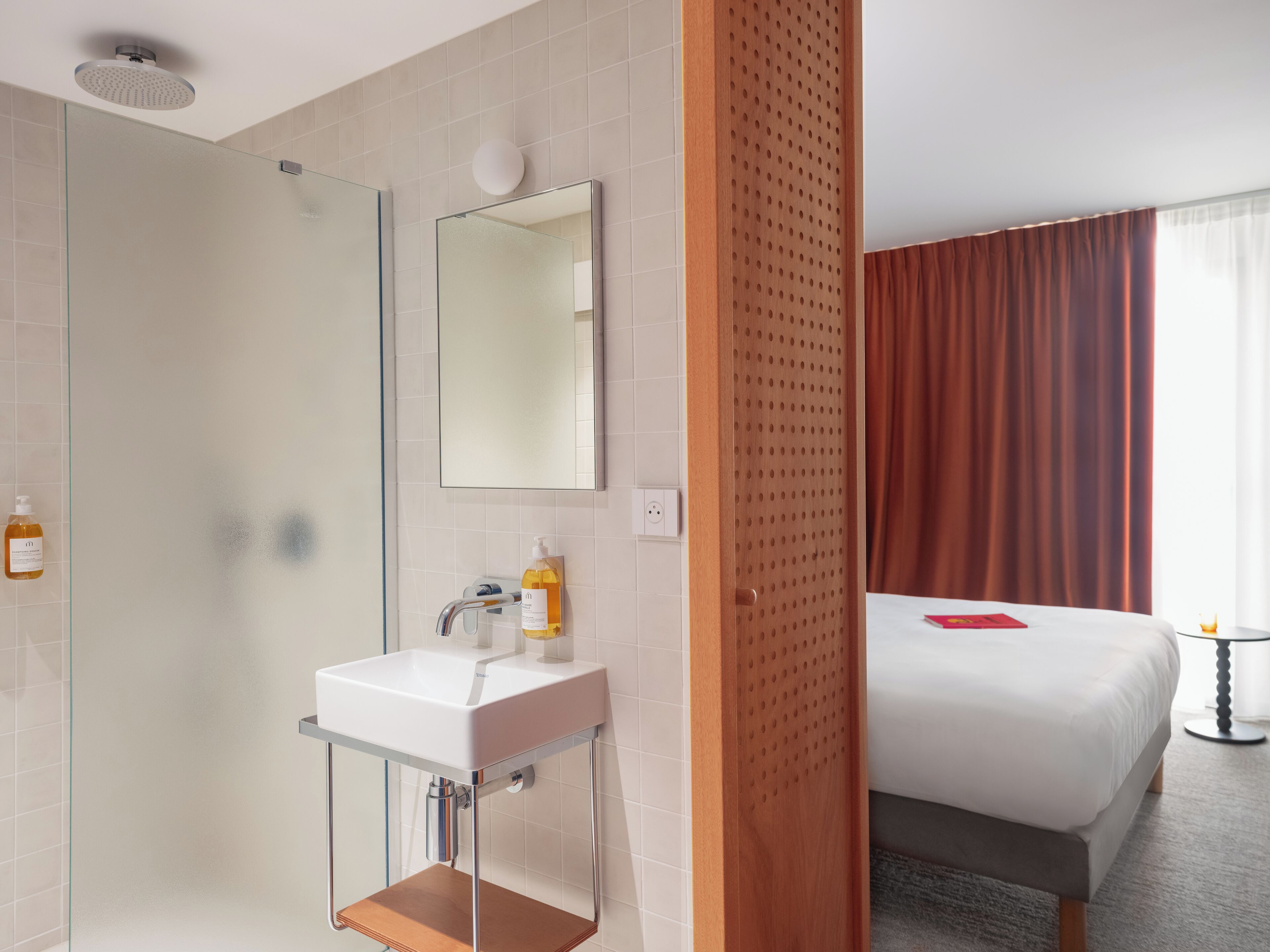 Habitación de hotel con ducha, lavabo, espejo, cama y ventana con cortinas.