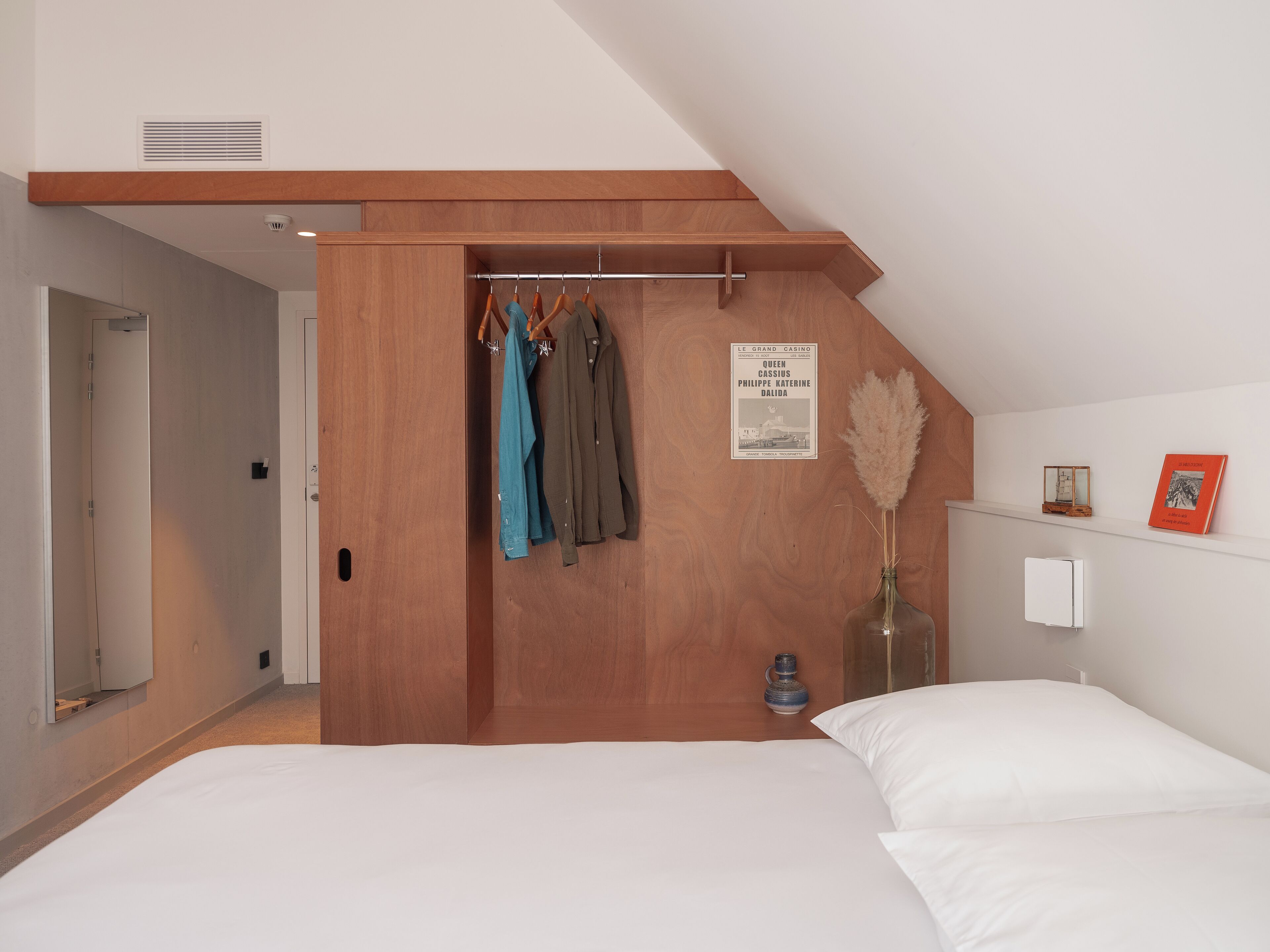 Une chambre avec une armoire en bois, un portant à vêtements, un lit avec une literie blanche, un vase contenant des plumes d’herbe des pampas et un tableau encadré accroché au mur.