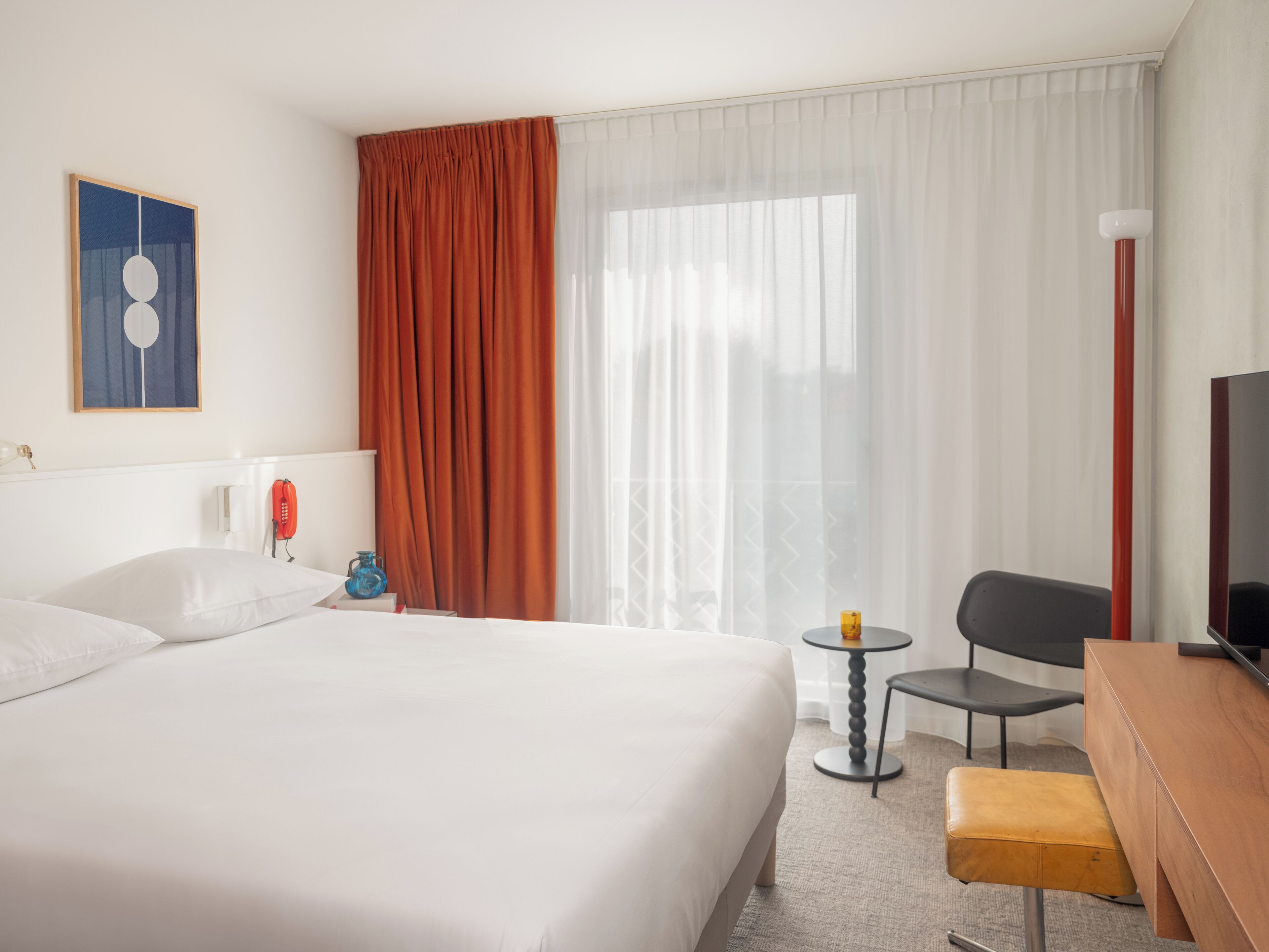 A hotel room with a large bed, a desk, a chair, a small table, a telephone, and a painting on the wall.