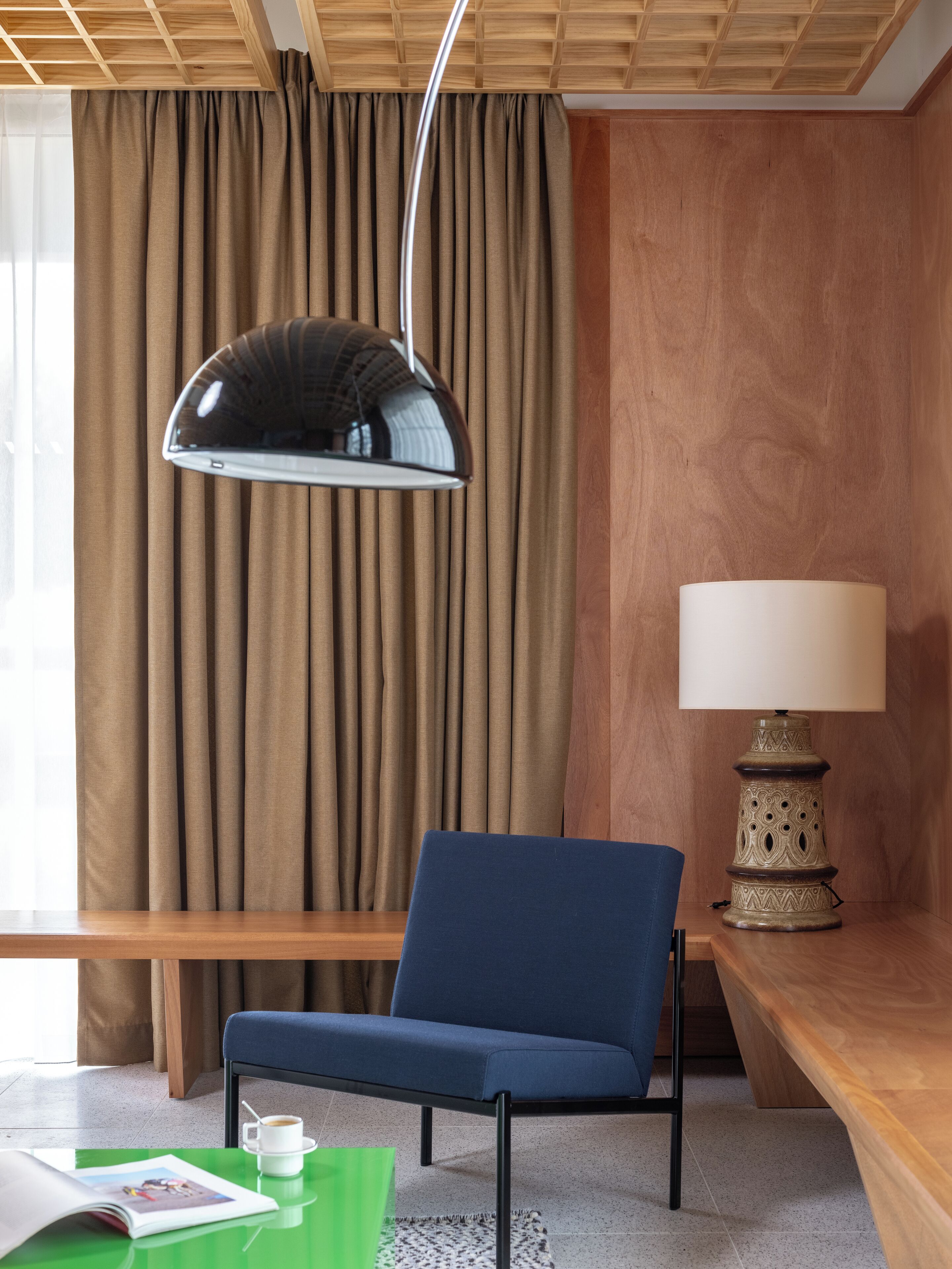 A modern room with a blue chair, a table with a cup and a magazine, a lamp, and a window with curtains.