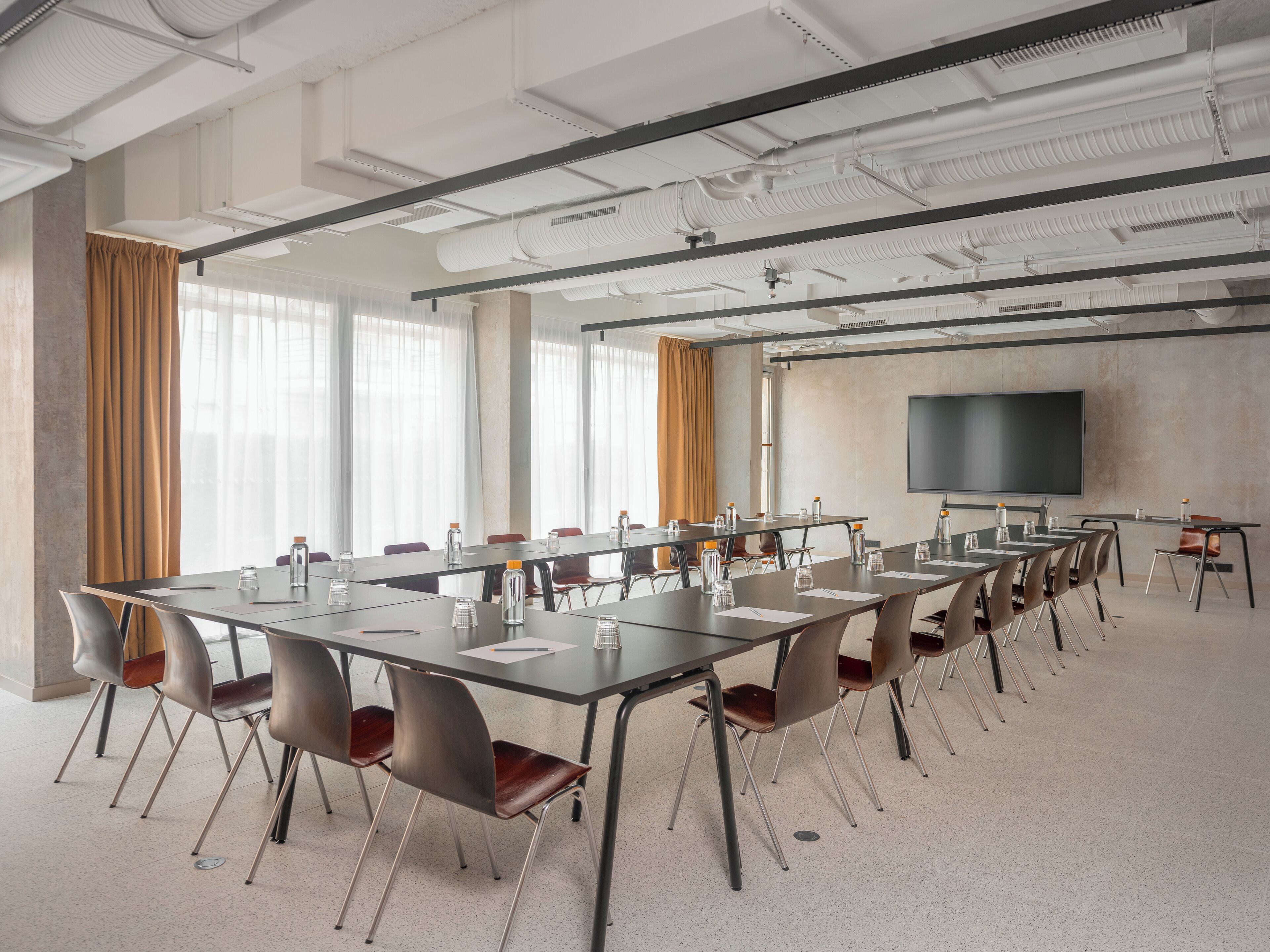 Un salón de conferencias con mesas largas, sillas, una pantalla de proyector y ventanas amplias con cortinas.