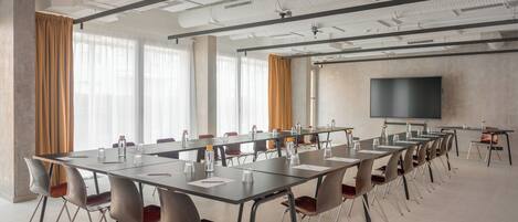 Un salón de conferencias con mesas largas, sillas, una pantalla de proyector y ventanas amplias con cortinas.