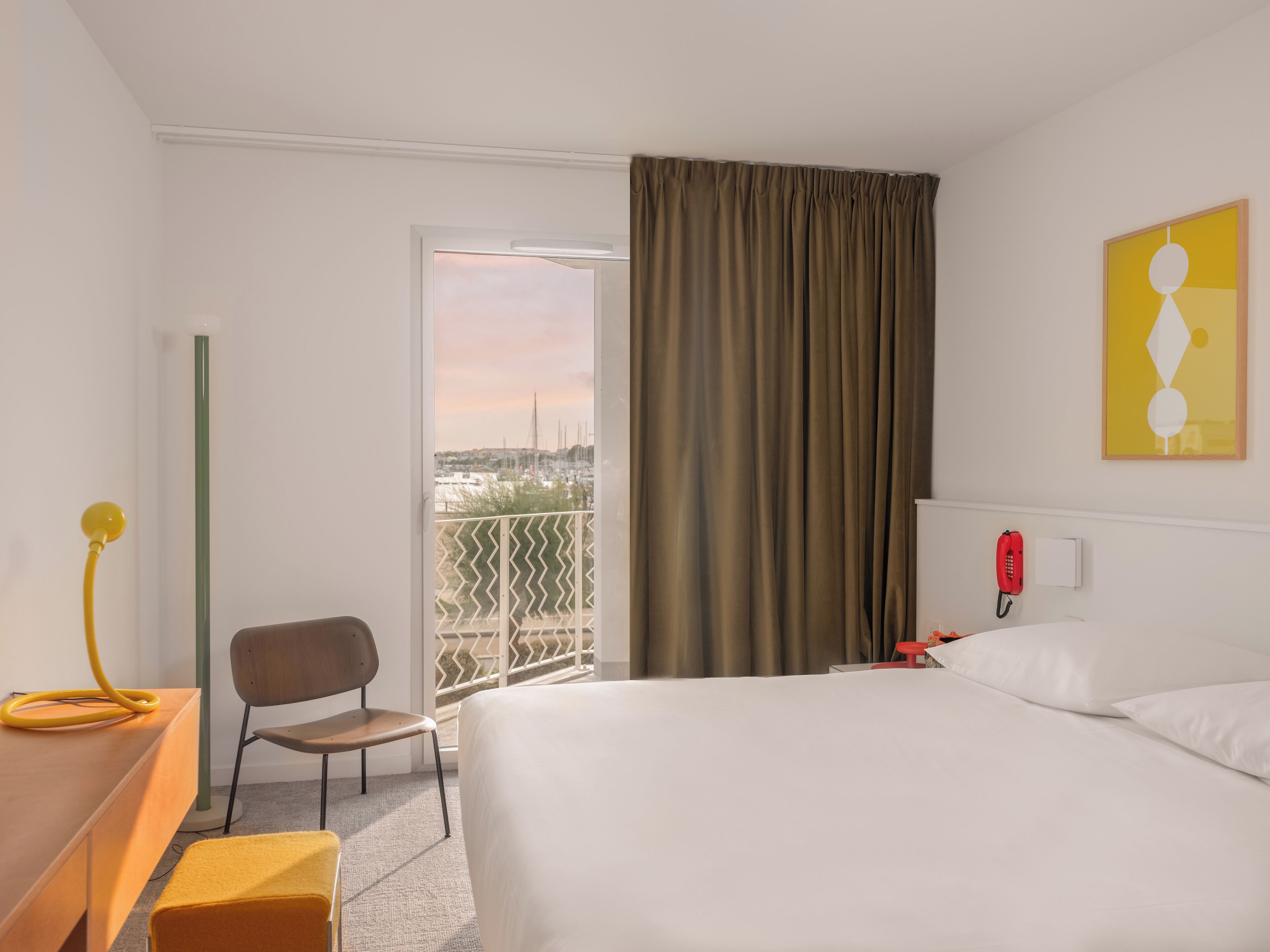 A hotel room with a bed, a desk, a chair, a telephone, and a view of the city through a window with curtains.
