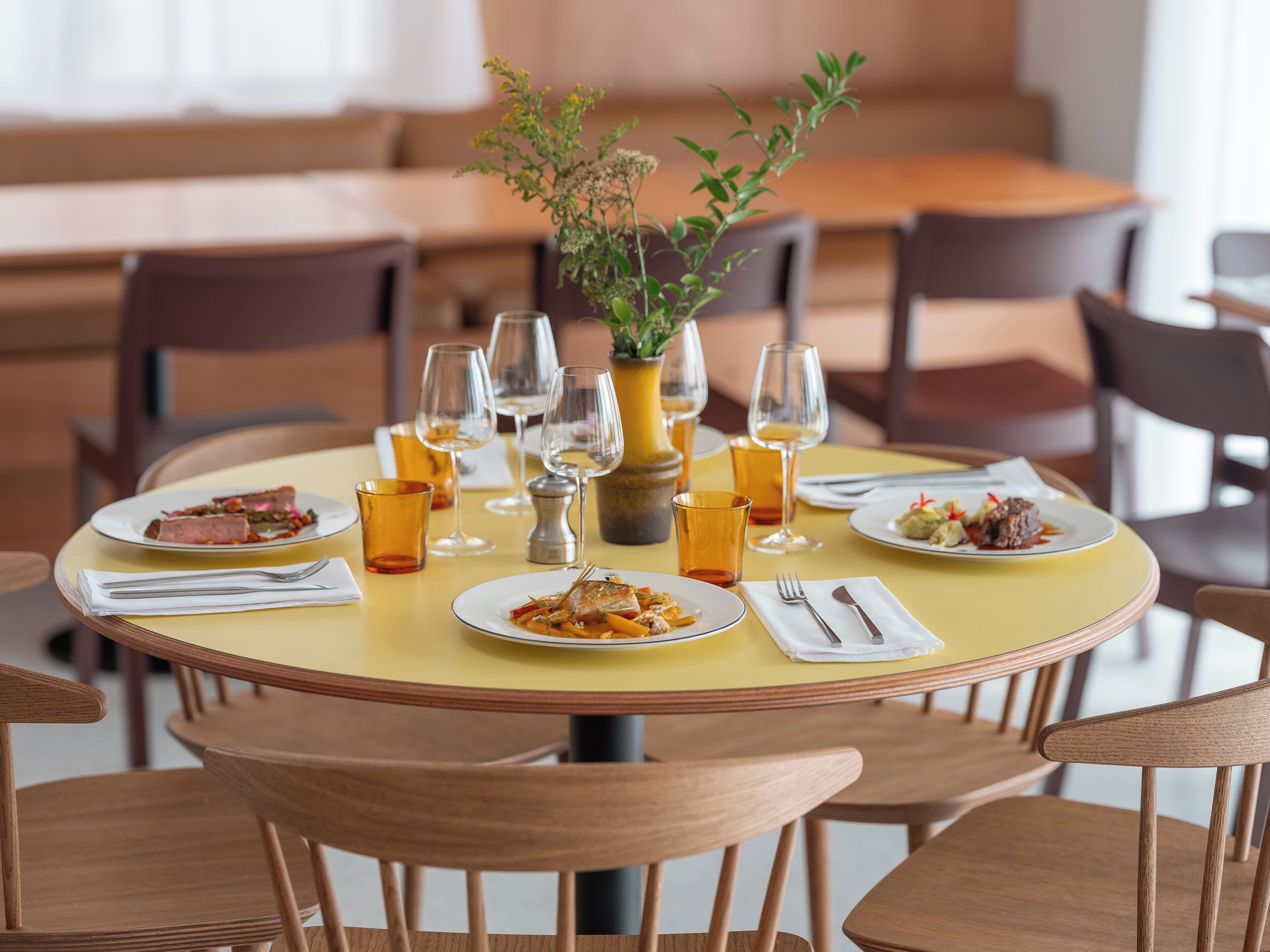 Una mesa redonda de comedor con mantel amarillo, rodeada de sillas de madera. Hay platos de comida, copas de vino y un florero con una planta sobre la mesa.