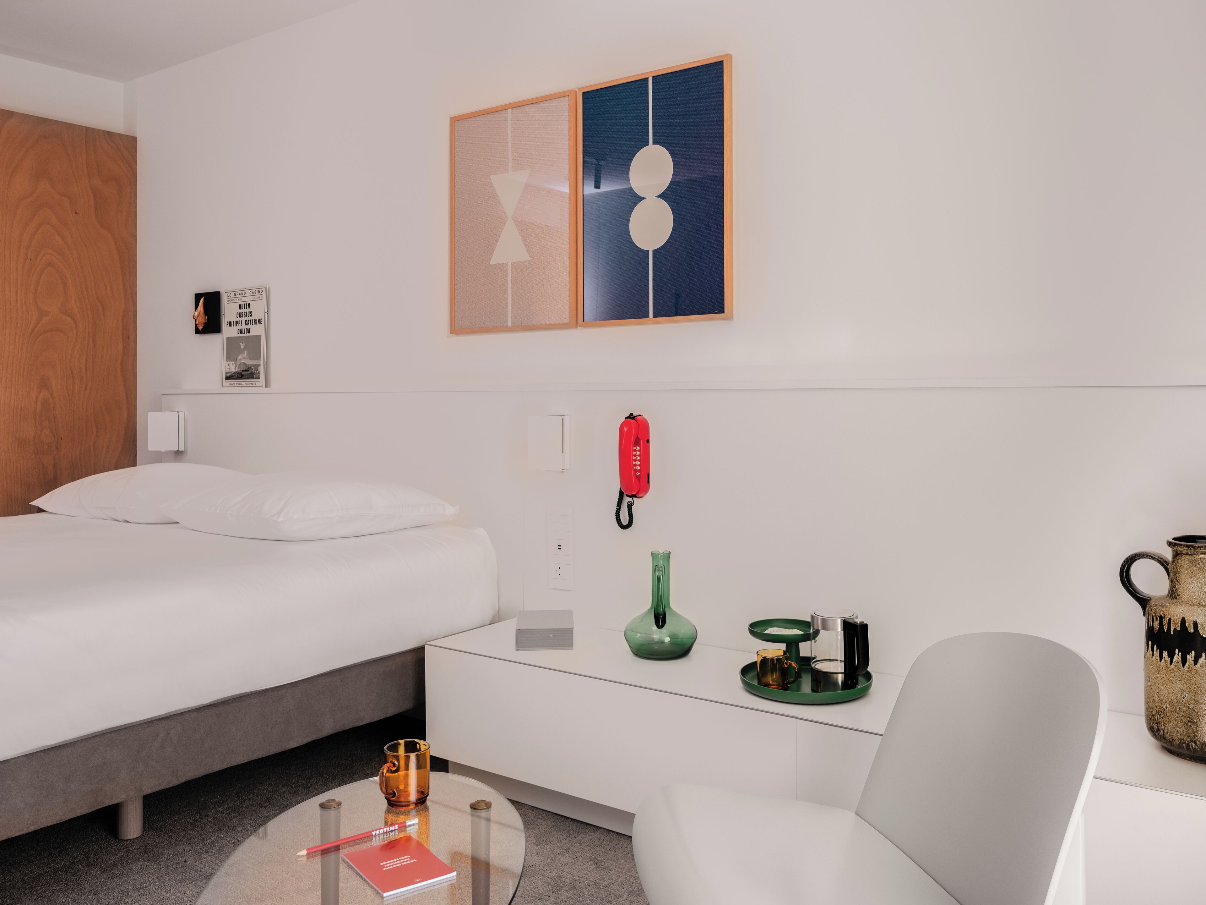 Ett hotellrum med en säng, ett skrivbord med en telefon, en stol och ett soffbord med en bok och en mugg.