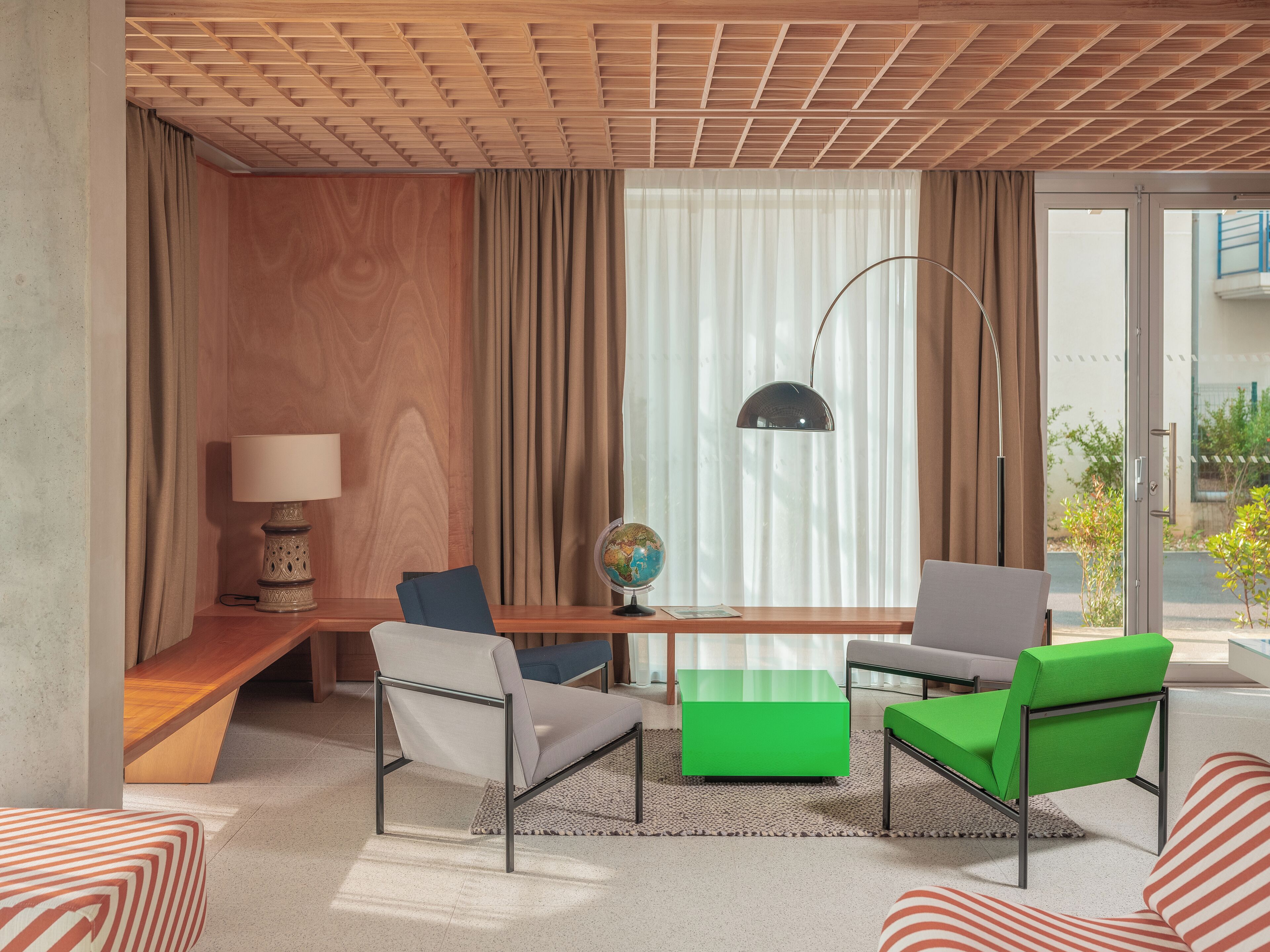 Une chambre d’hôtel moderne avec un plafond en bois, un bureau sur lequel se trouve un globe terrestre, deux chaises, une lampe et une vue sur l’extérieur.