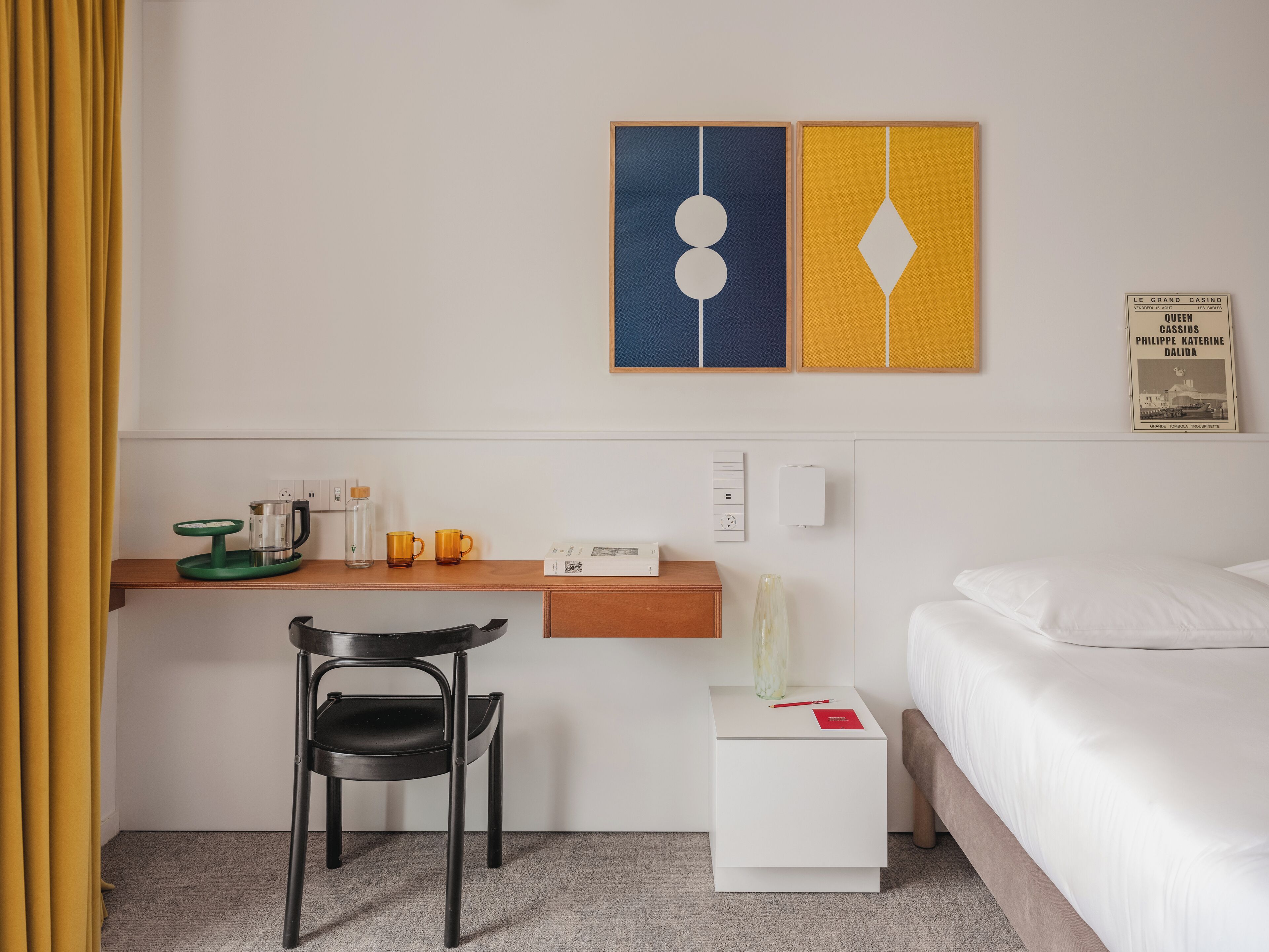 A hotel room with a bed, a desk with a chair, a small table with a coffee maker and cups, and a wall with two framed pictures.