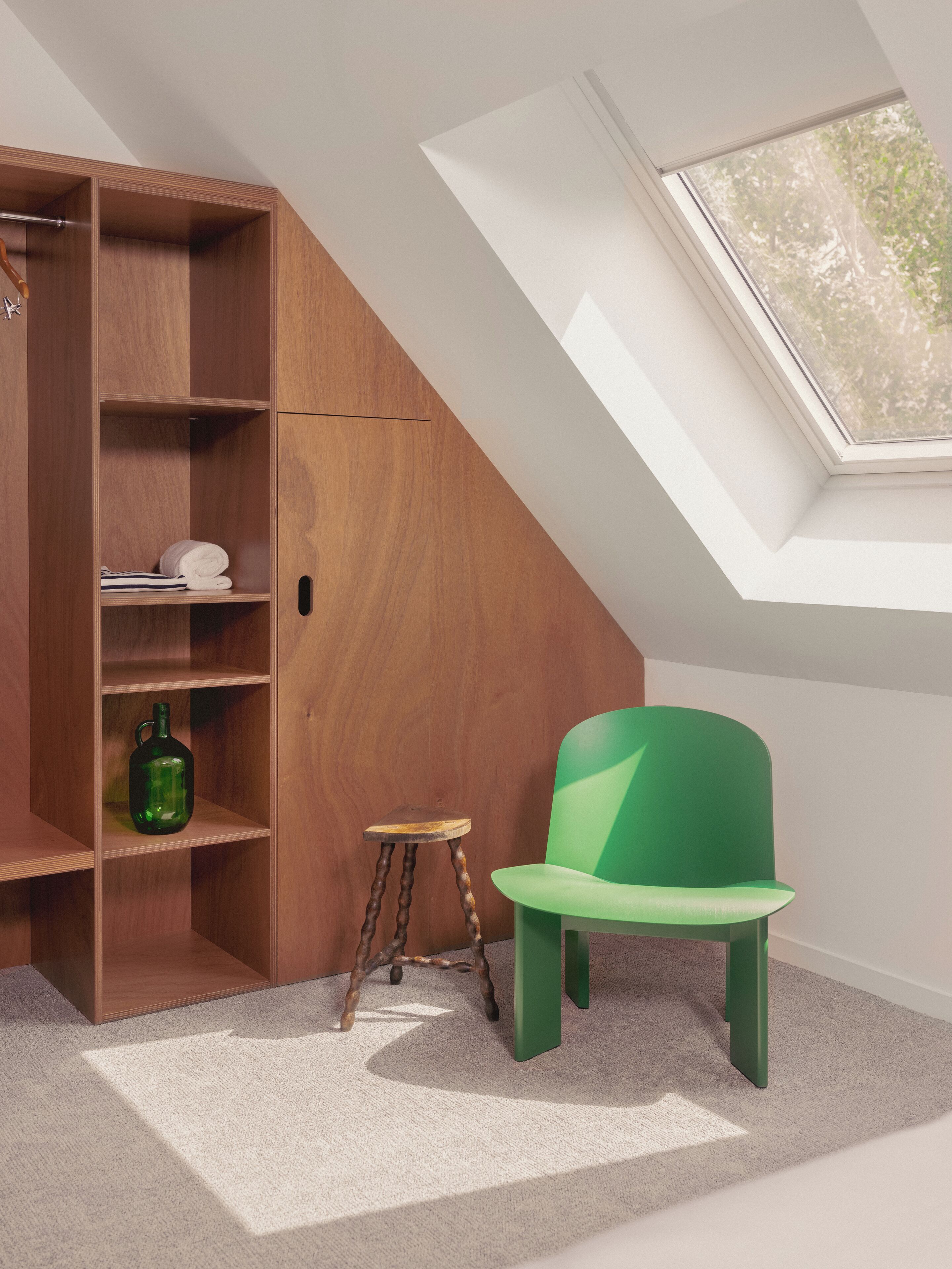 A room with a sloped ceiling, a skylight, a wooden shelving unit, a green chair, and a small stool.