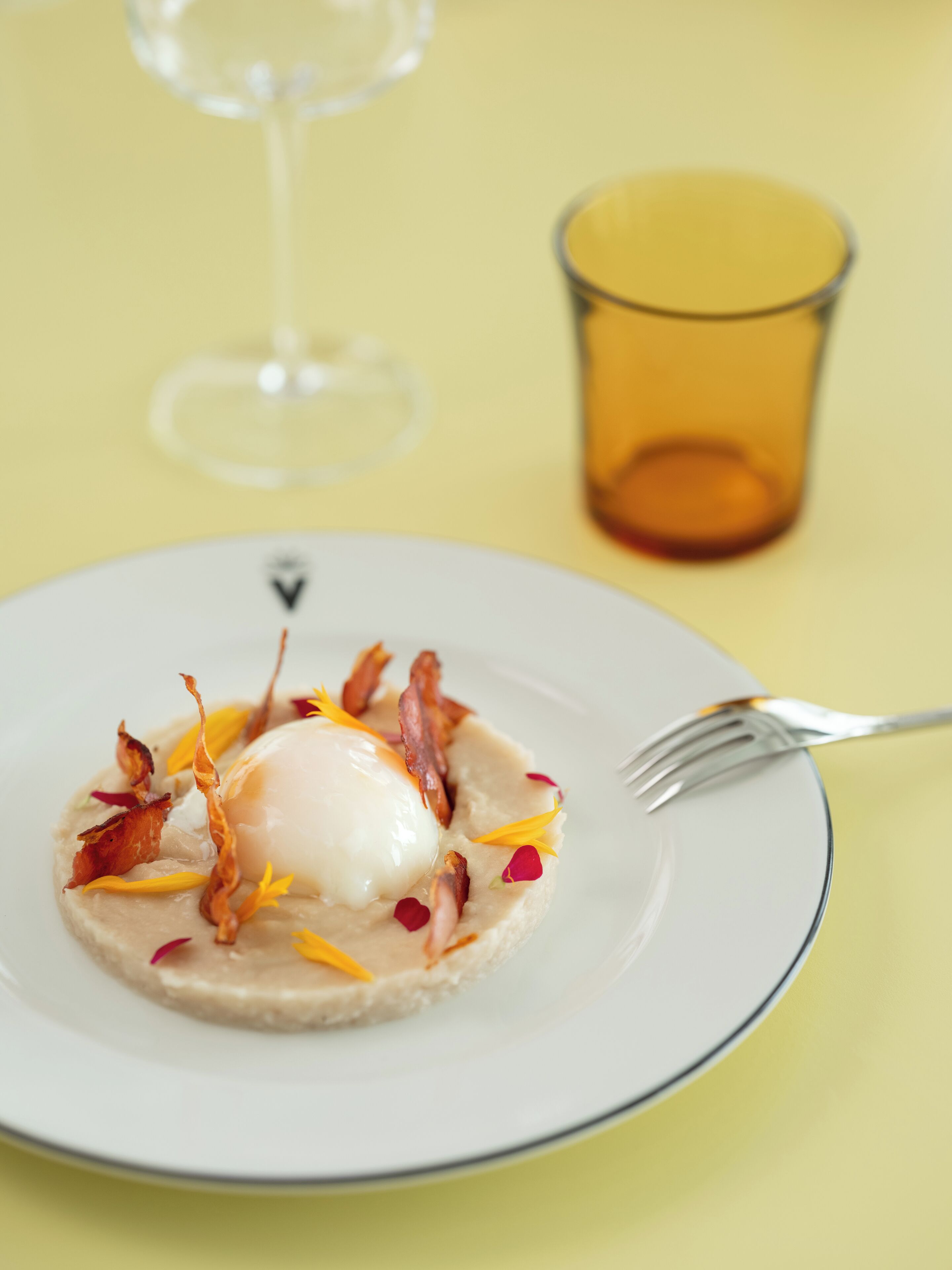 Une assiette blanche bordée de bleu, contenant un plat composé d’un œuf poché, de bacon et de quelques accompagnements.