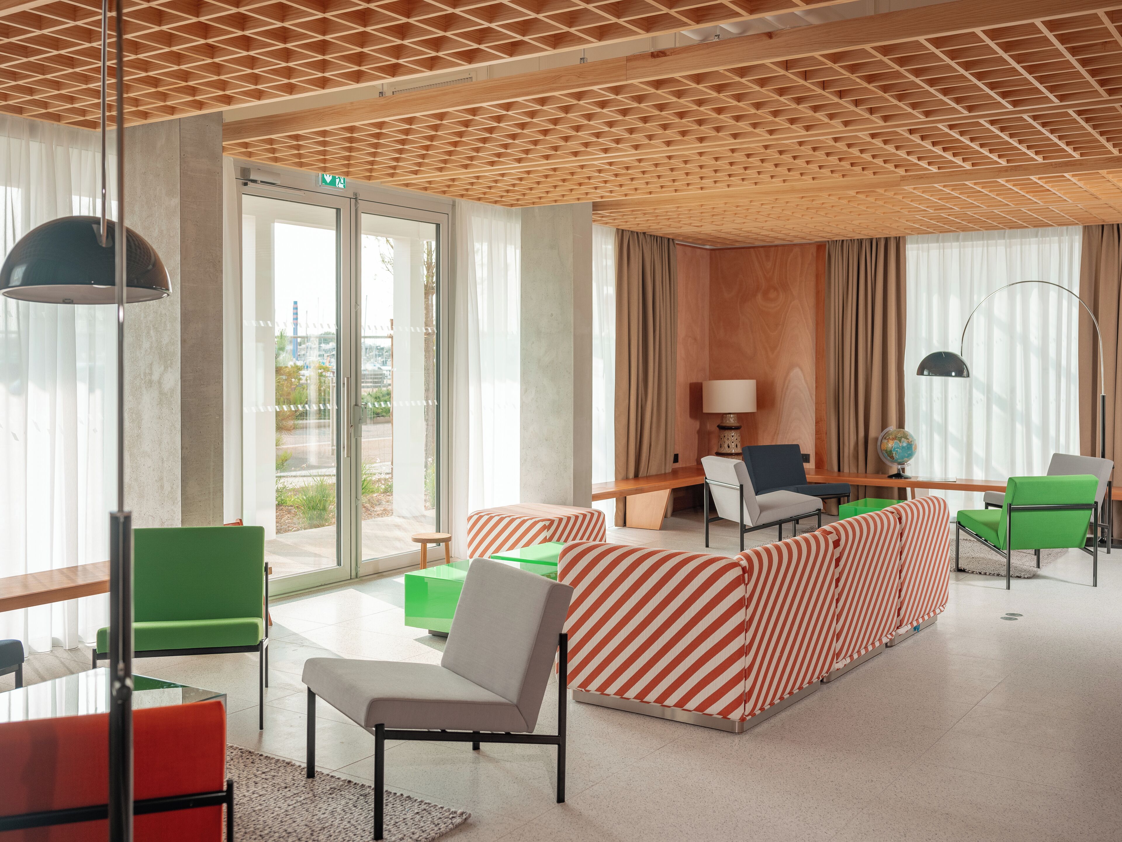 Un hall d’hôtel moderne avec un plafond en bois, de grandes fenêtres et un espace salon doté de canapés à rayures et de fauteuils verts.
