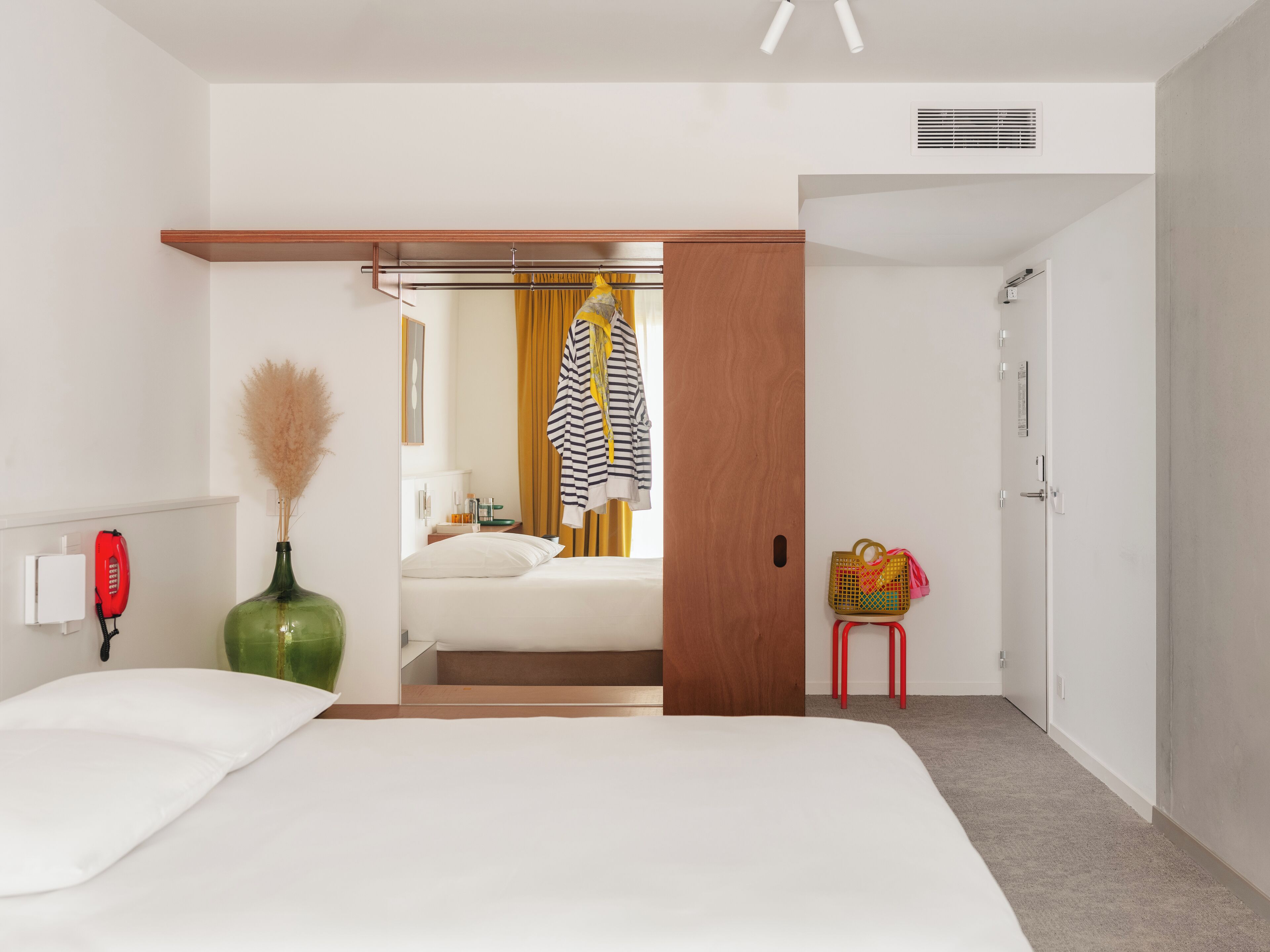 A hotel room with a bed, a wardrobe, a mirror, a red telephone, a green vase, and a small red stool.