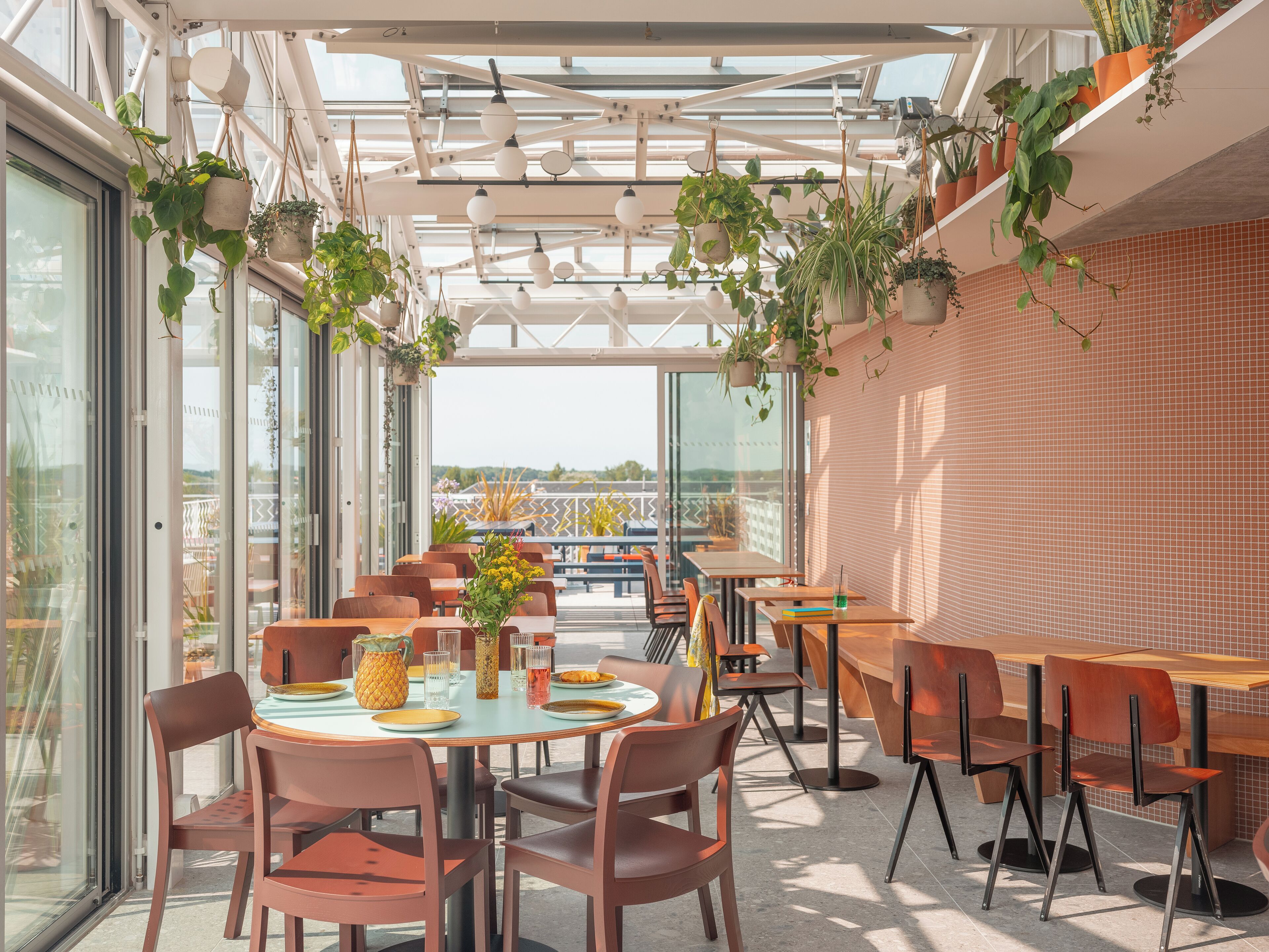 A modern outdoor dining area with round tables and chairs, surrounded by glass walls and potted plants.