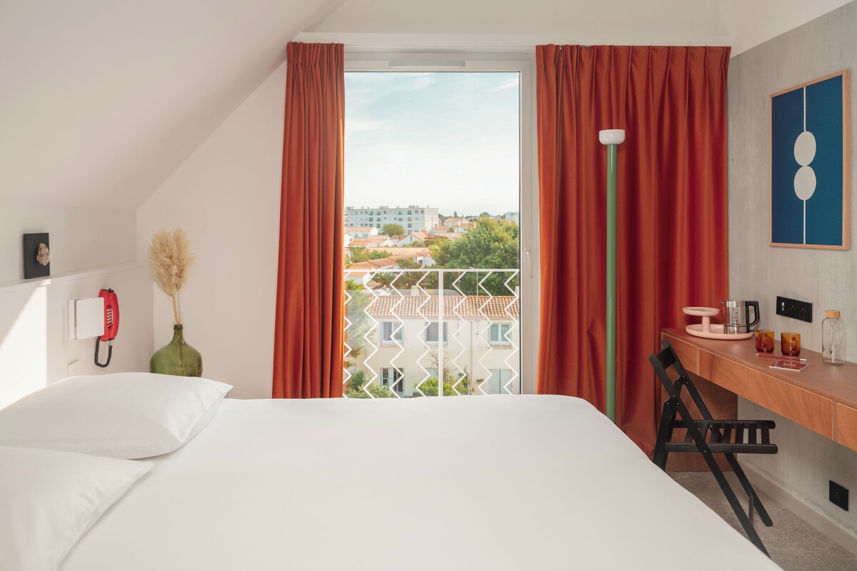 A hotel room with a bed, a desk, a chair, a phone, a vase with pampas grass, and a view of buildings through a window with red curtains.