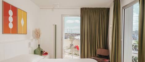Een hotelkamer met een bed, een stoel, een tafel met een vaas en uitzicht op de stad door een groot raam.