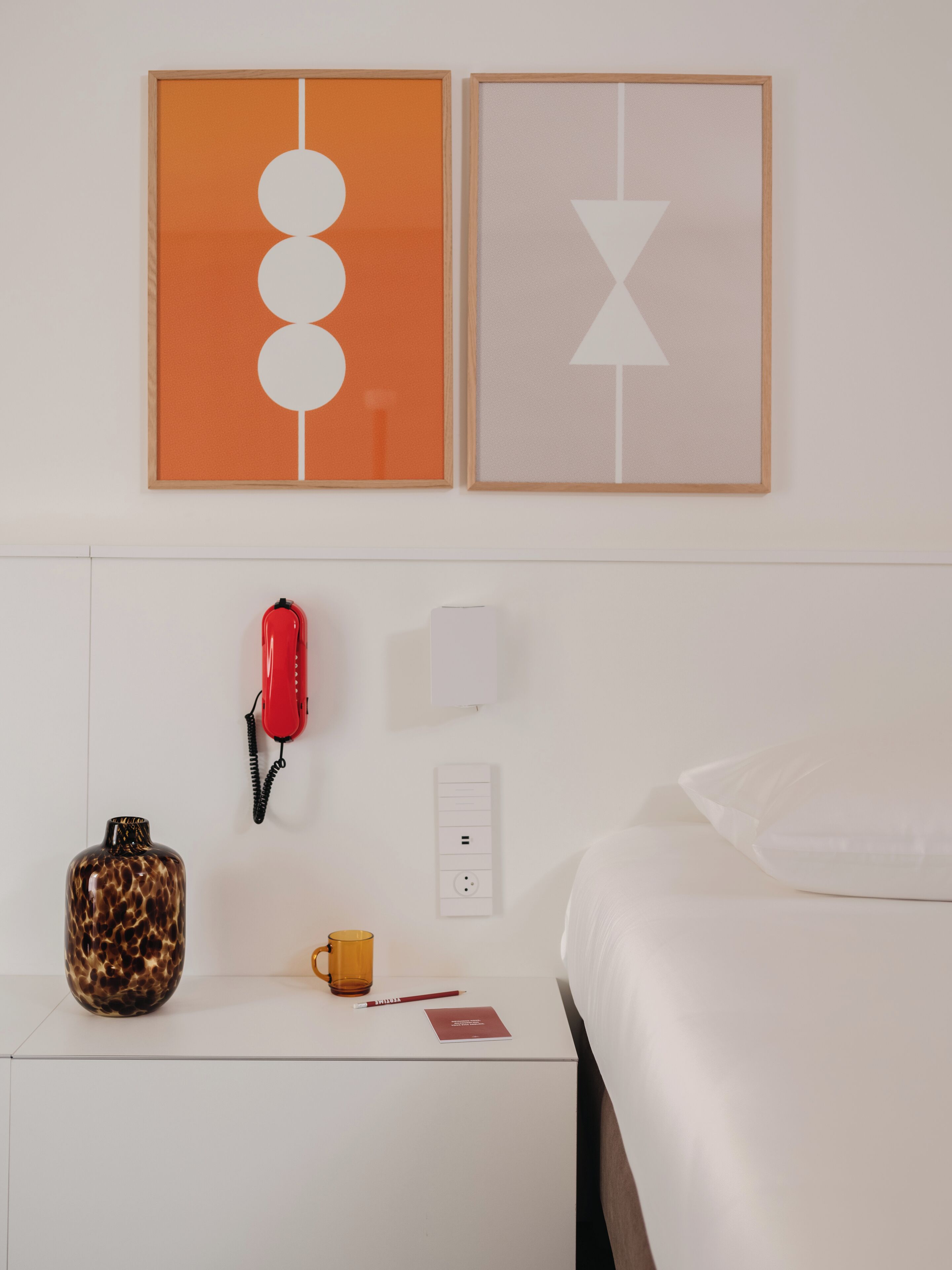 A hotel room with a bed, a nightstand with a red telephone, a vase, a mug, and a notepad.