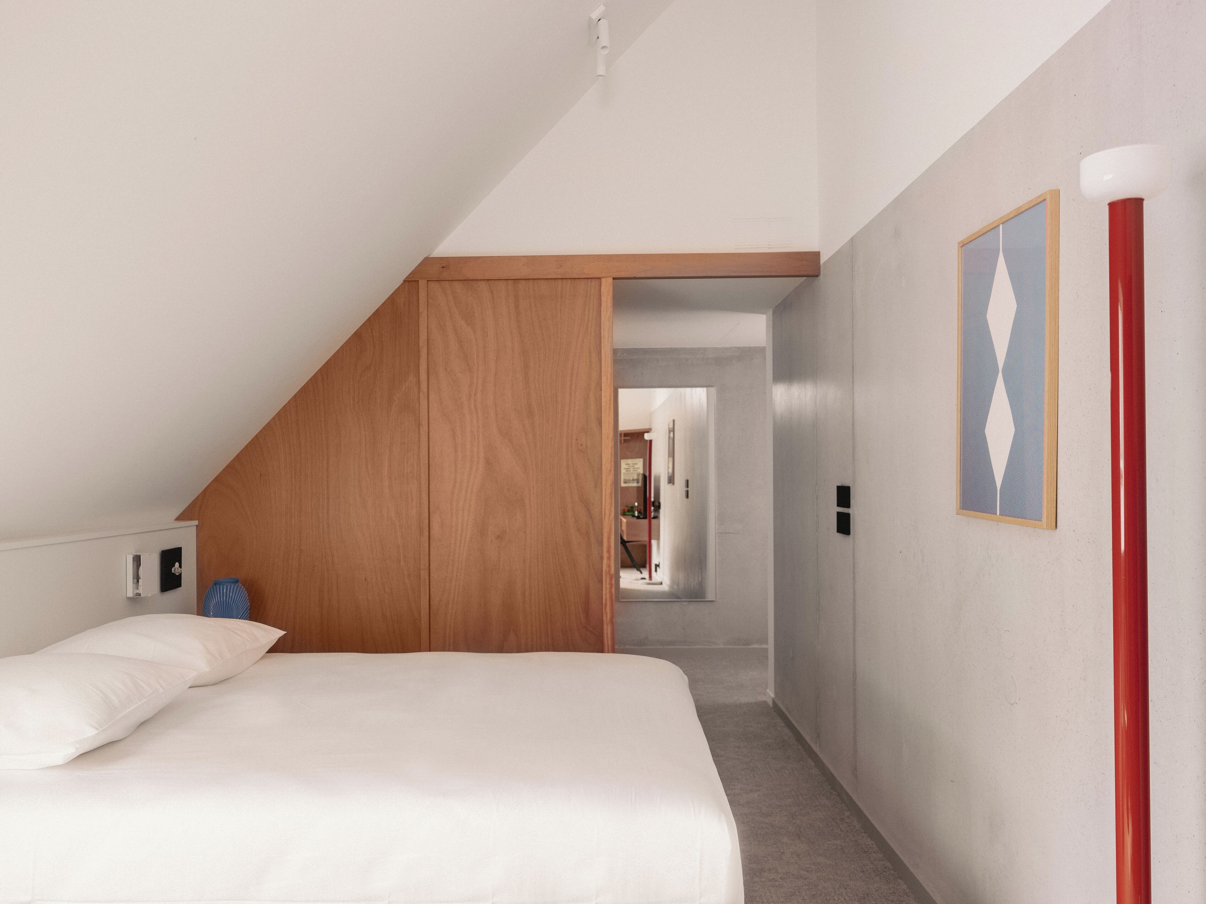 Ein Schlafzimmer mit schrägen Wänden, einem Bett mit weißer Bettwäsche, einem Holzschrank und einem Flur, der zu einem Badezimmer führt.
