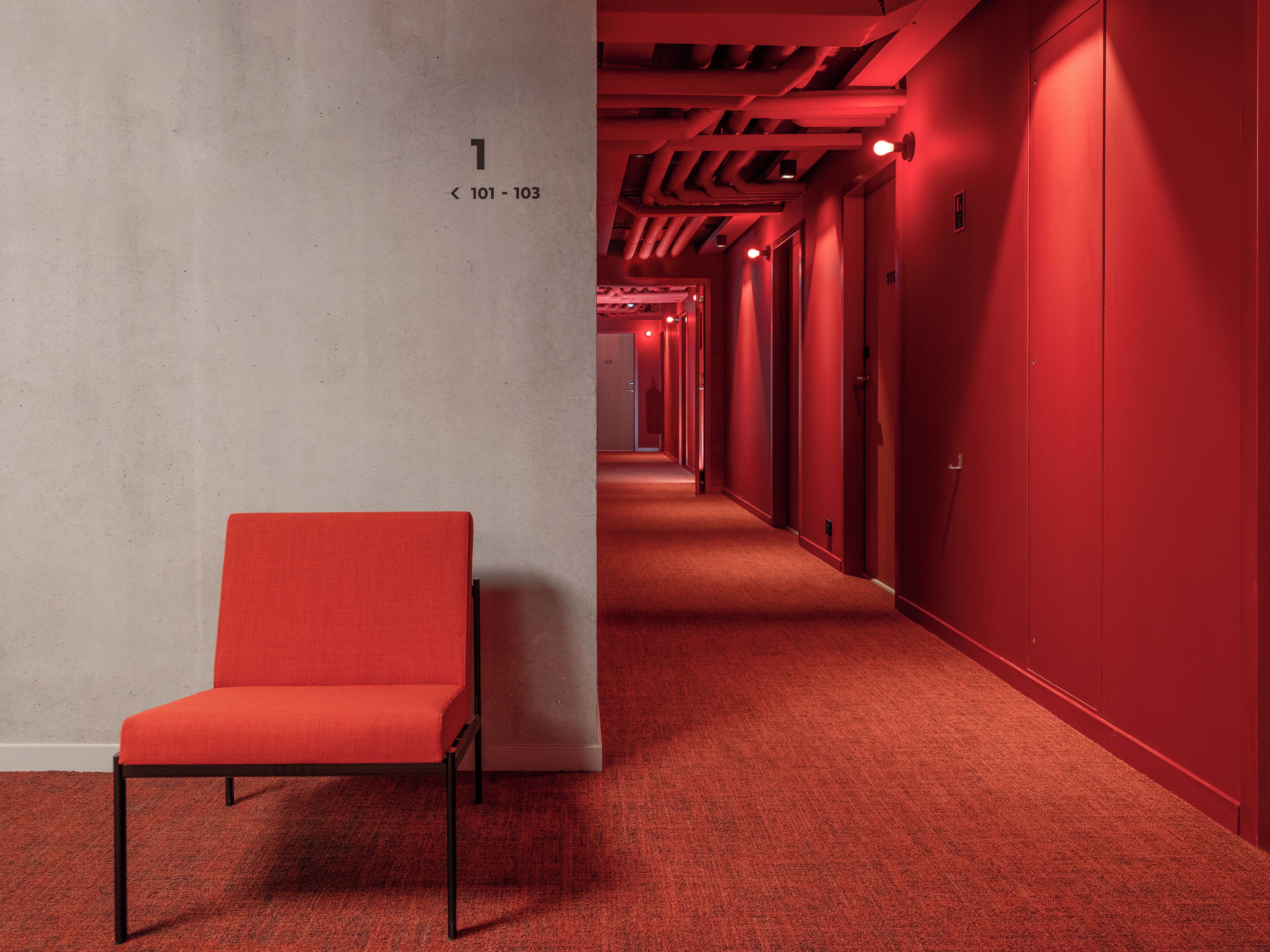 Un pasillo con alfombra roja, una silla roja, puertas numeradas y un pilar de concreto.