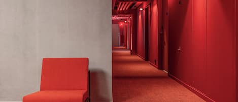 Un pasillo con alfombra roja, una silla roja, puertas numeradas y un pilar de concreto.