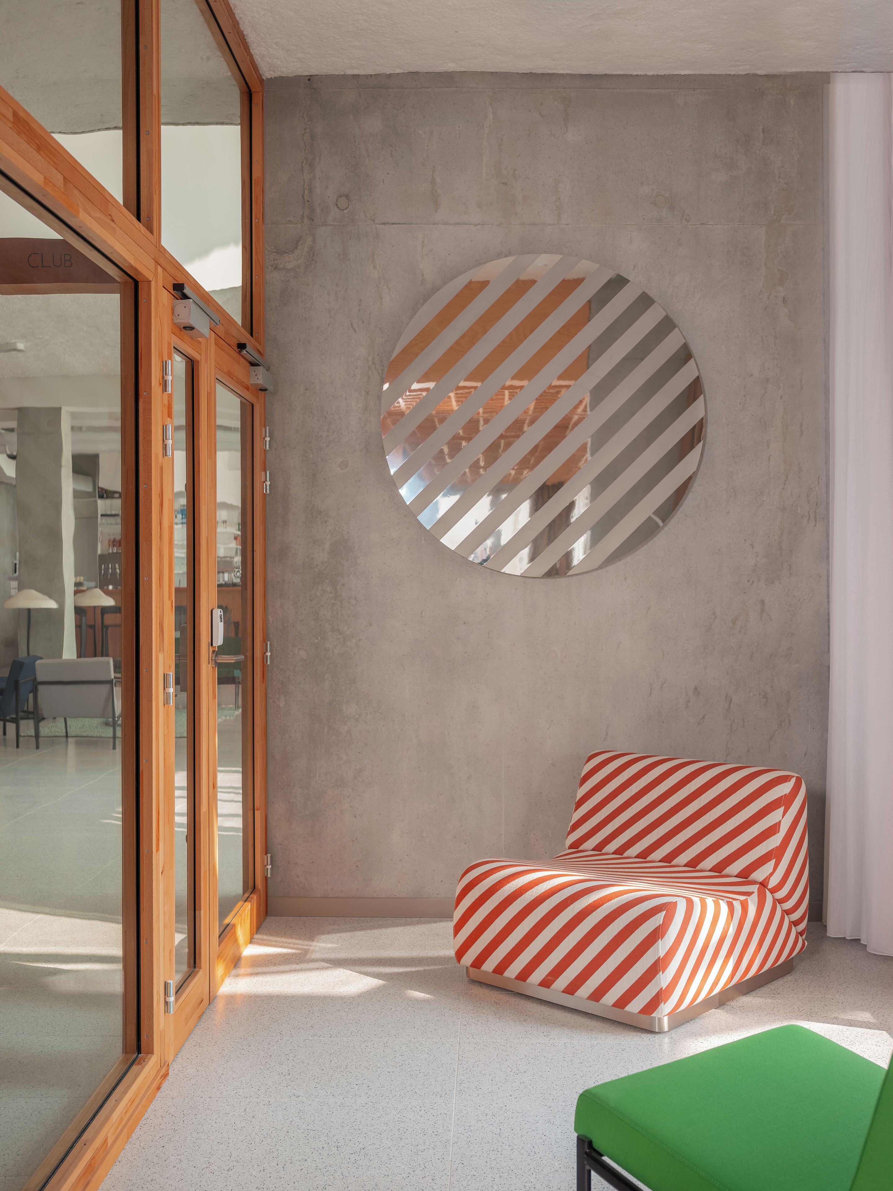 Una habitación moderna con un ventanal grande, un espejo redondo con rayas diagonales y sillas tapizadas a rayas rojas y blancas.