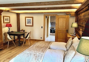 A bedroom with a bed, a wooden table, chairs, a lamp, and a door leading to a kitchen.