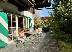 Une terrasse dallée de pierres, avec une table et des chaises, un banc doté d’un coussin, et un bâtiment aux ornements et aux fenêtres de couleur verte.