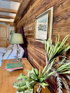 A wooden wall with a framed picture, a bedside table with books and a lamp, and a plant.
