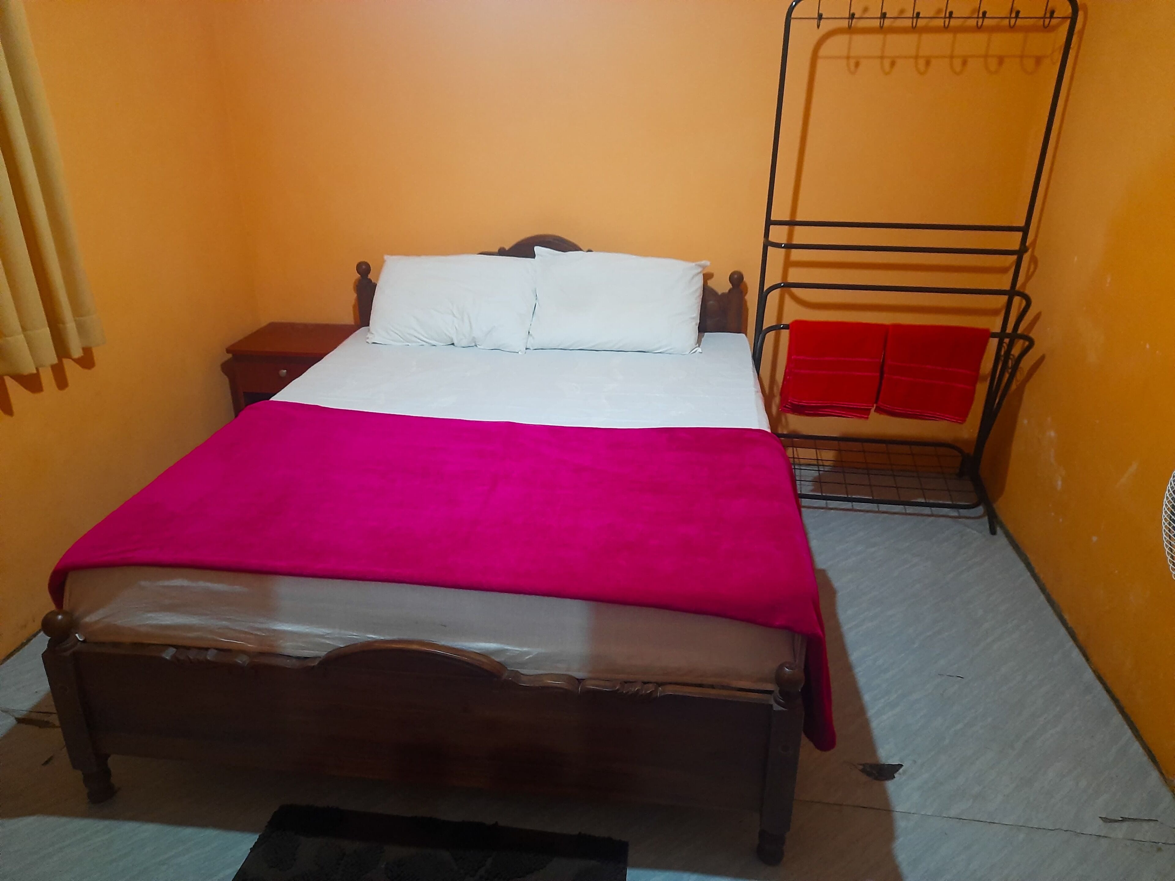 Una cama individual con estructura de madera, un colchón blanco y una manta rosa. Junto a la cama hay una mesita de noche de madera con una lámpara. Una escalera metálica está fijada a la pared en el lado derecho de la cama.