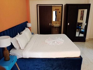 A hotel room with a large bed, a nightstand with a lamp, a bathroom with a sink and mirror, and an orange wall. - Abode Bungalow 05 (Assagao)