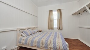 A bedroom with a wooden bed, a window with curtains, a small bedside table, and a wall-mounted shelf with hangers.