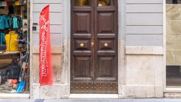 A building with a large wooden double door, a visible storefront with clothing items, and a red flag with white text. - Hiro Studio - Spanish Steps (Roma)