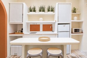 A compact kitchenette with a white countertop, two stools, a microwave, a refrigerator, and potted plants on shelves.