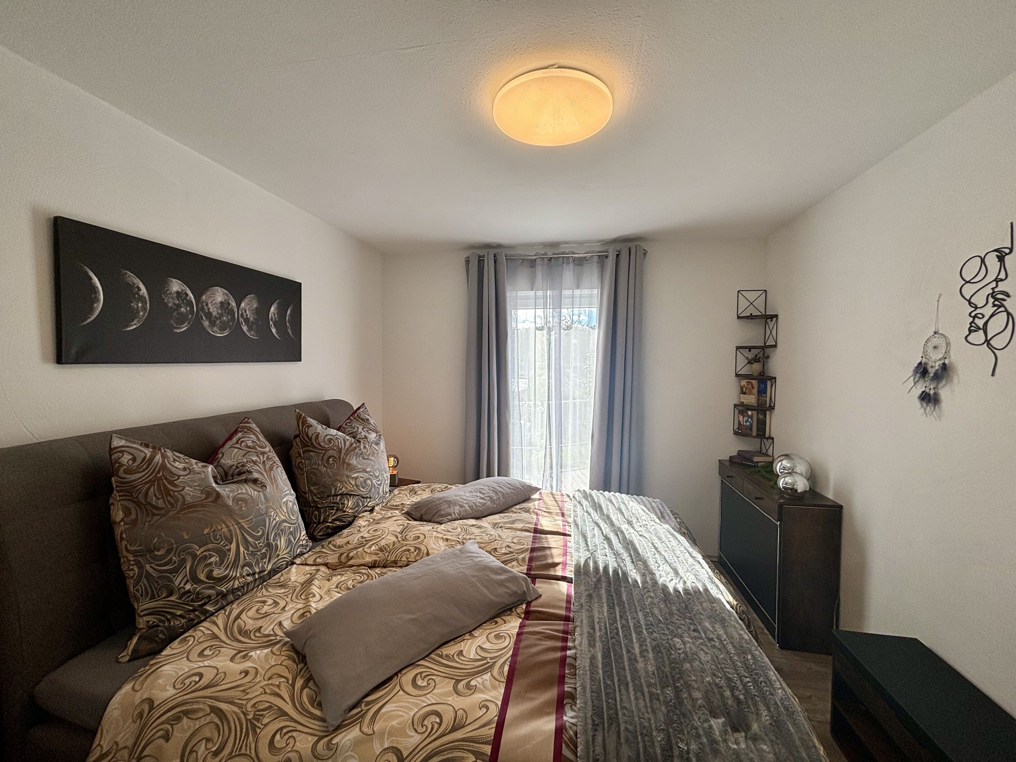 Une chambre à coucher comprenant un lit, une table de chevet, une œuvre d’art accrochée au mur, une fenêtre avec des rideaux et un luminaire au plafond.