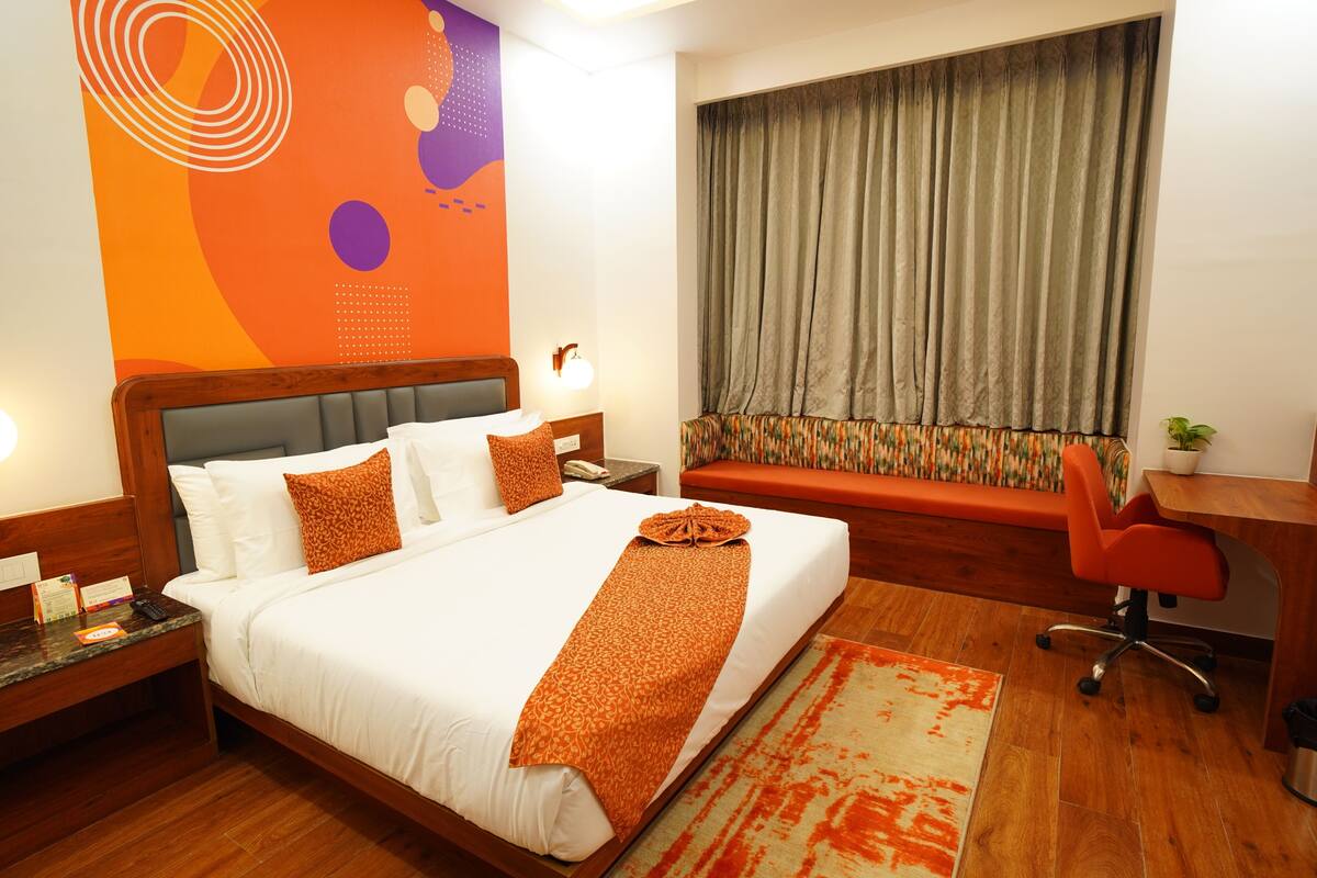 A hotel room with a large bed, a desk, a chair, a sofa, a large window with curtains, and a wall with abstract art.