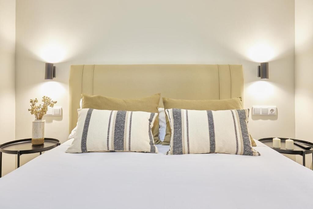 Una cama bien hecha con dos almohadas, un jarrón con flores secas y dos lámparas de pared a cada lado.