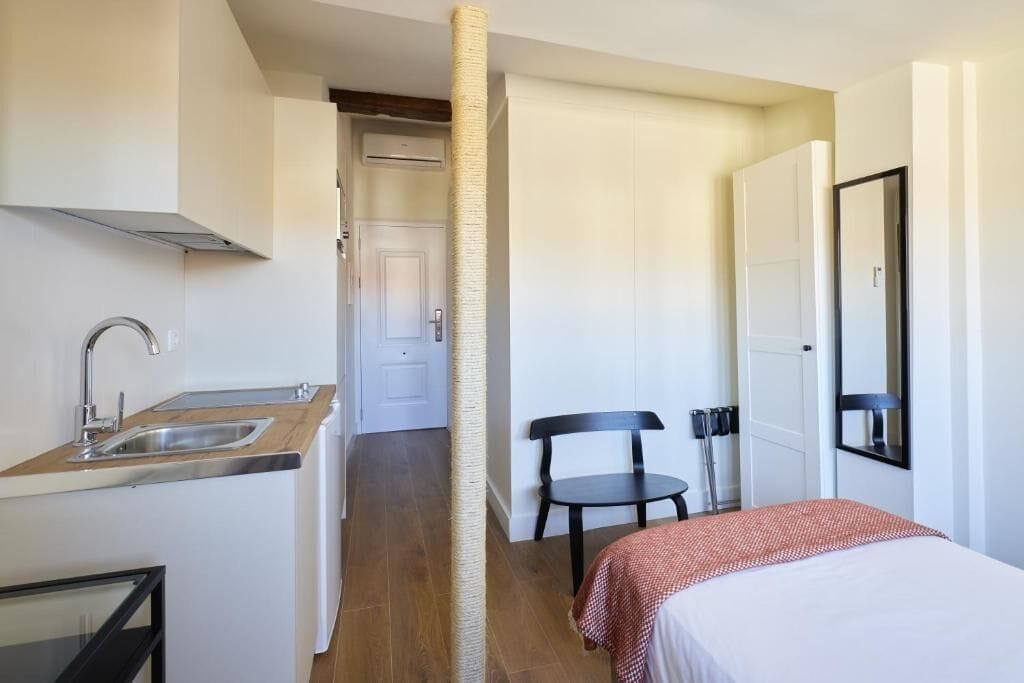 A compact hotel room with a kitchenette, a bed, a chair, and a door leading to a bathroom.