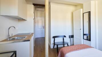 A compact hotel room with a kitchenette, a bed, a chair, and a door leading to a bathroom.