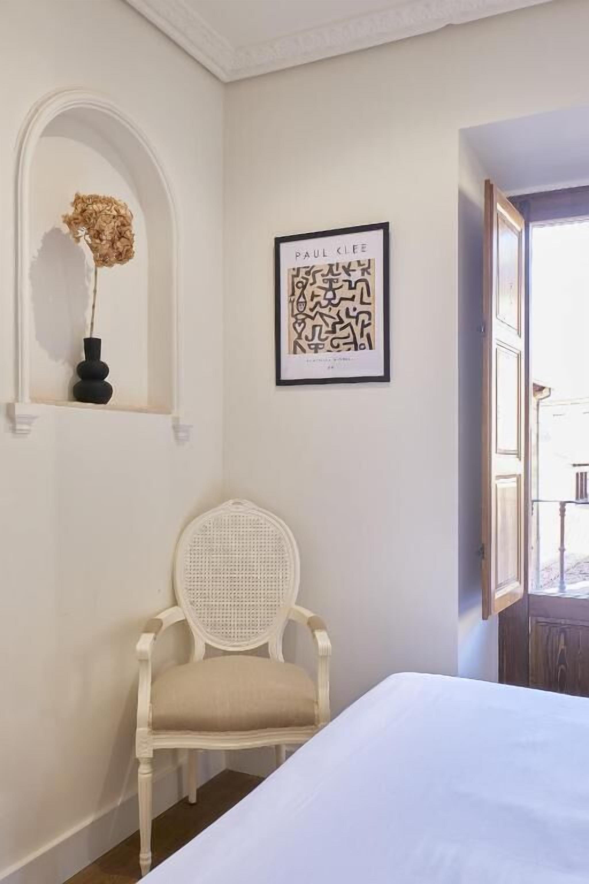 A room with a bed, a white chair, a framed artwork, and a vase with dried flowers on a shelf.
