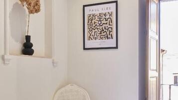 A room with a bed, a white chair, a framed artwork, and a vase with dried flowers on a shelf.