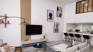 Una sala de estar moderna con un sofá blanco, una mesa de centro de vidrio y un televisor. Cuenta con una zona de cocina con taburetes de bar y una mesa de comedor.