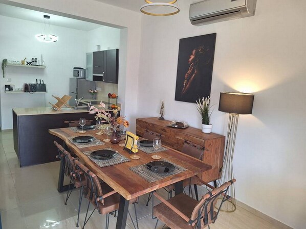 Un comedor moderno con una mesa de madera dispuesta para cuatro personas, rodeada de sillas. Se observa una cocina al fondo con un microondas y armarios.