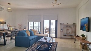 A spacious living room with a blue sofa, a coffee table, and a dining area with a table and chairs. - Paramount Gardens Resorts D204 (Tersefanou)