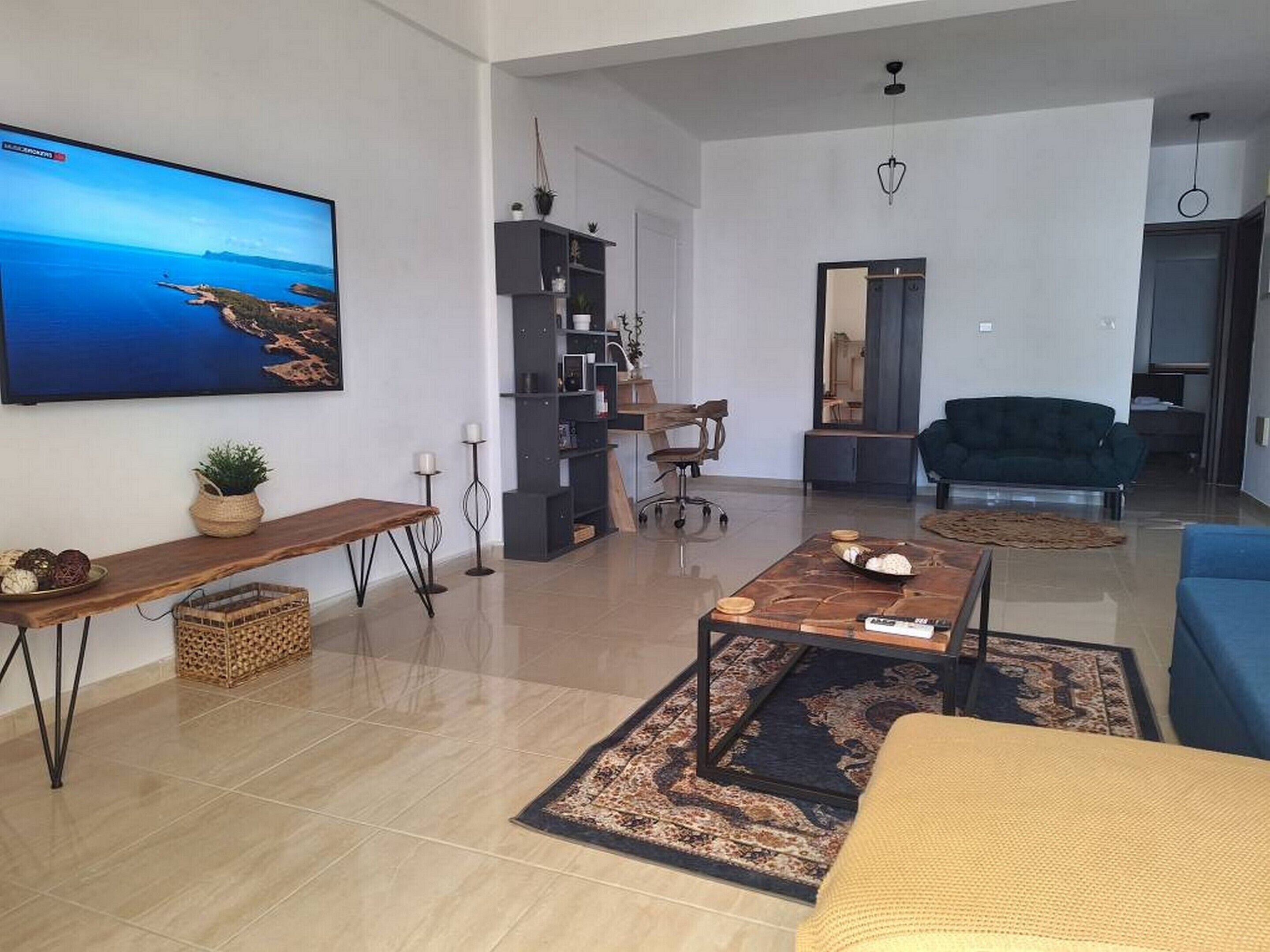Ett modernt vardagsrum med en platt-TV, ett soffbord i trä, en blå soffa och en grå soffa. Det finns också en bokhylla och ett skrivbord med en stol.