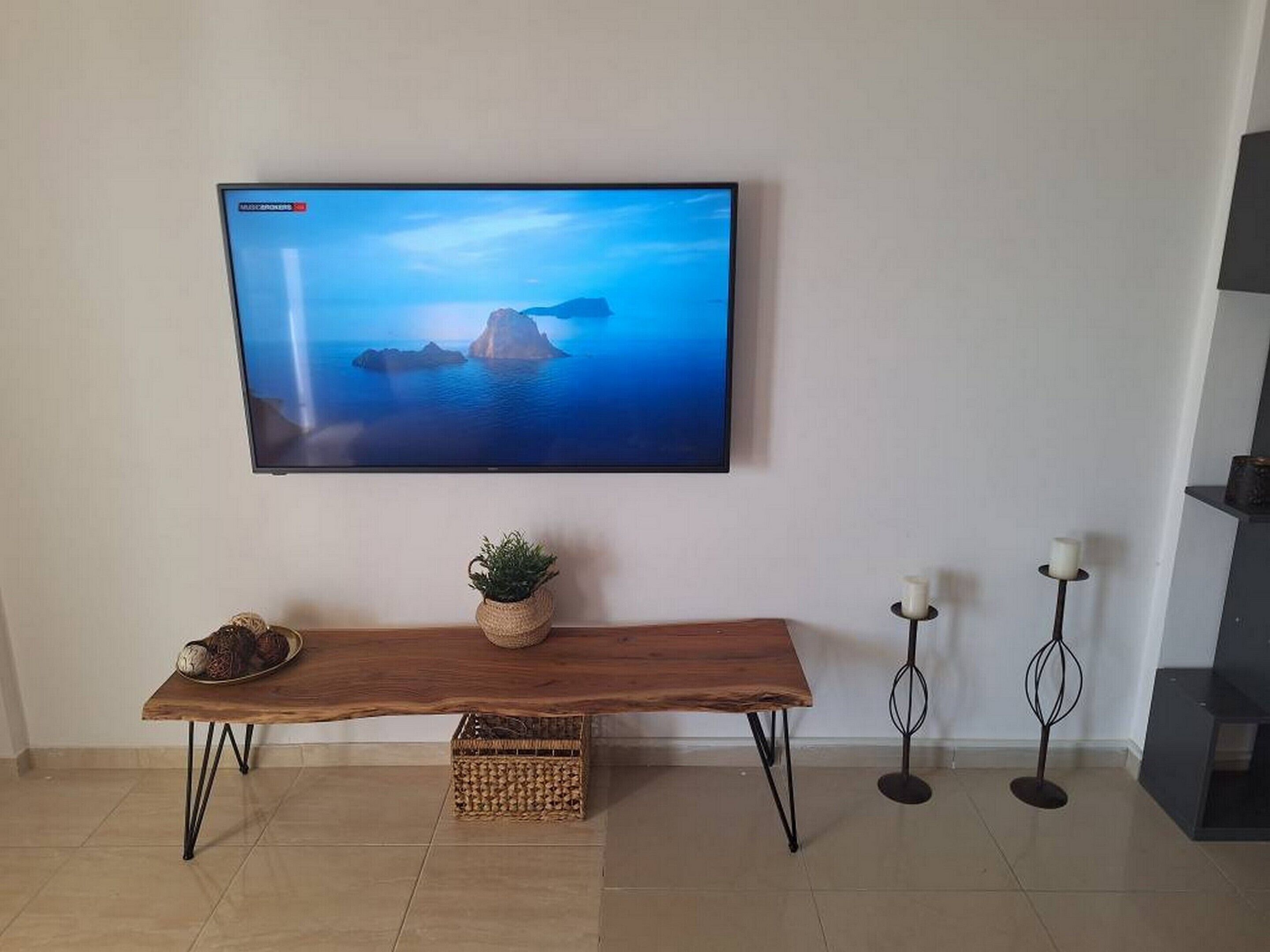 A flat-screen TV mounted on a wall displaying a scenic ocean view. Below the TV, there's a wooden bench with a woven basket and a small potted plant.
